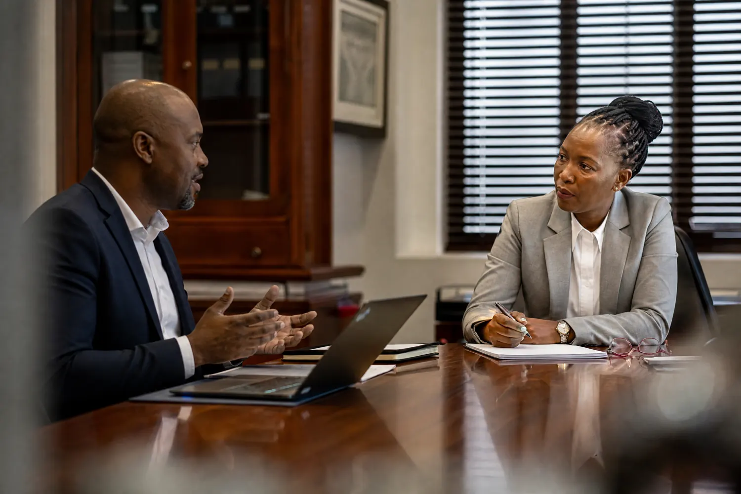 Two colleagues in a plain office discuss a document across a desk.
