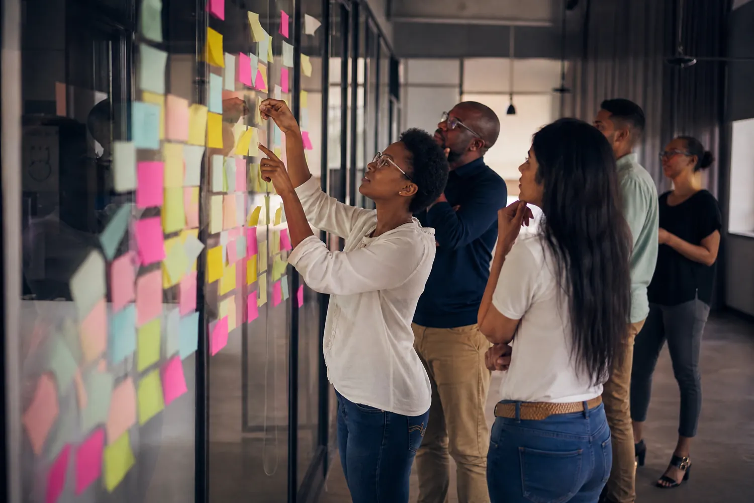 A team workshop, one person adding a sticky note to a glass wall already covered in notes while colleagues look on.