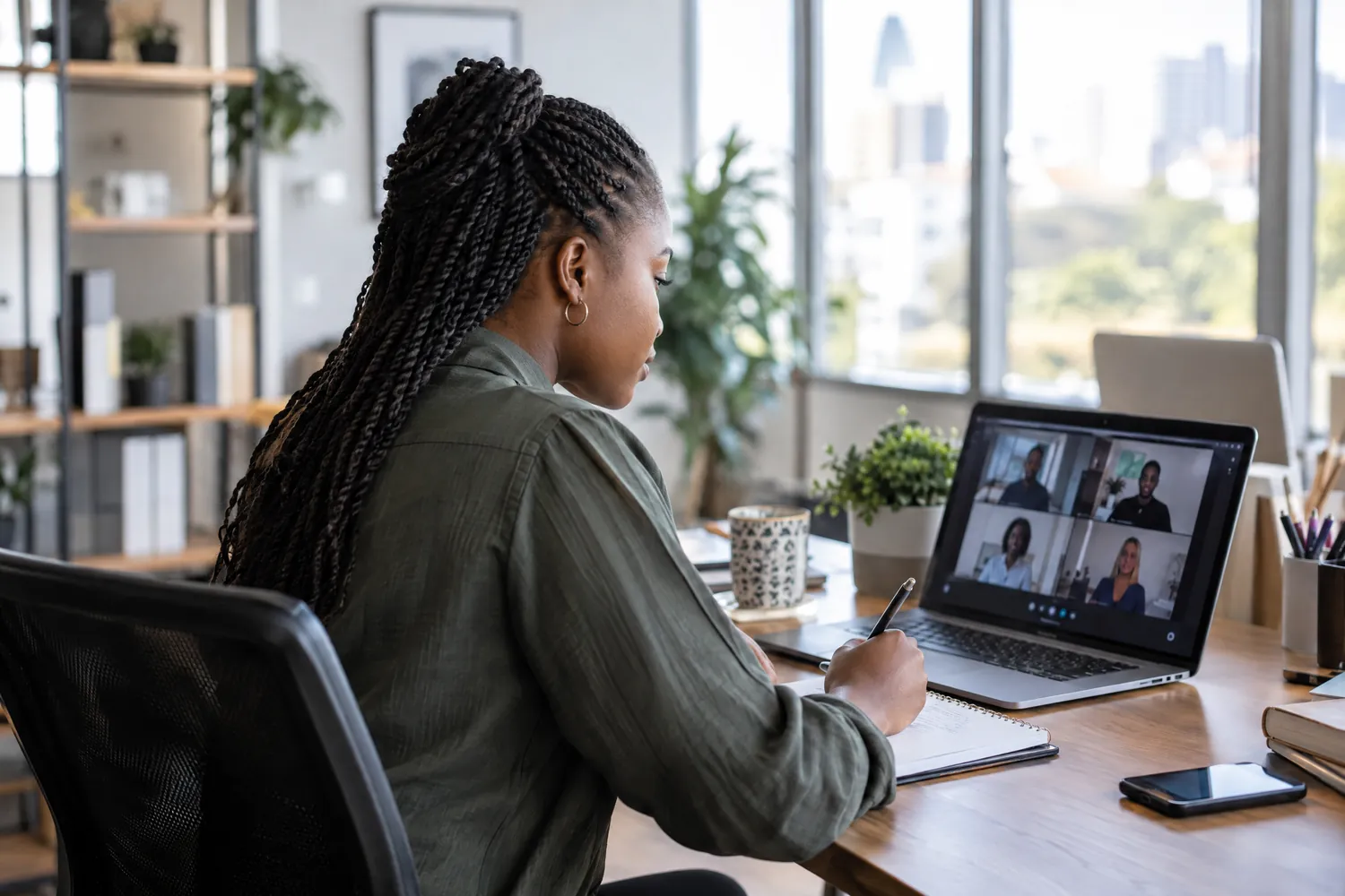 A team leader takes notes at her desk during a live video session on her laptop.