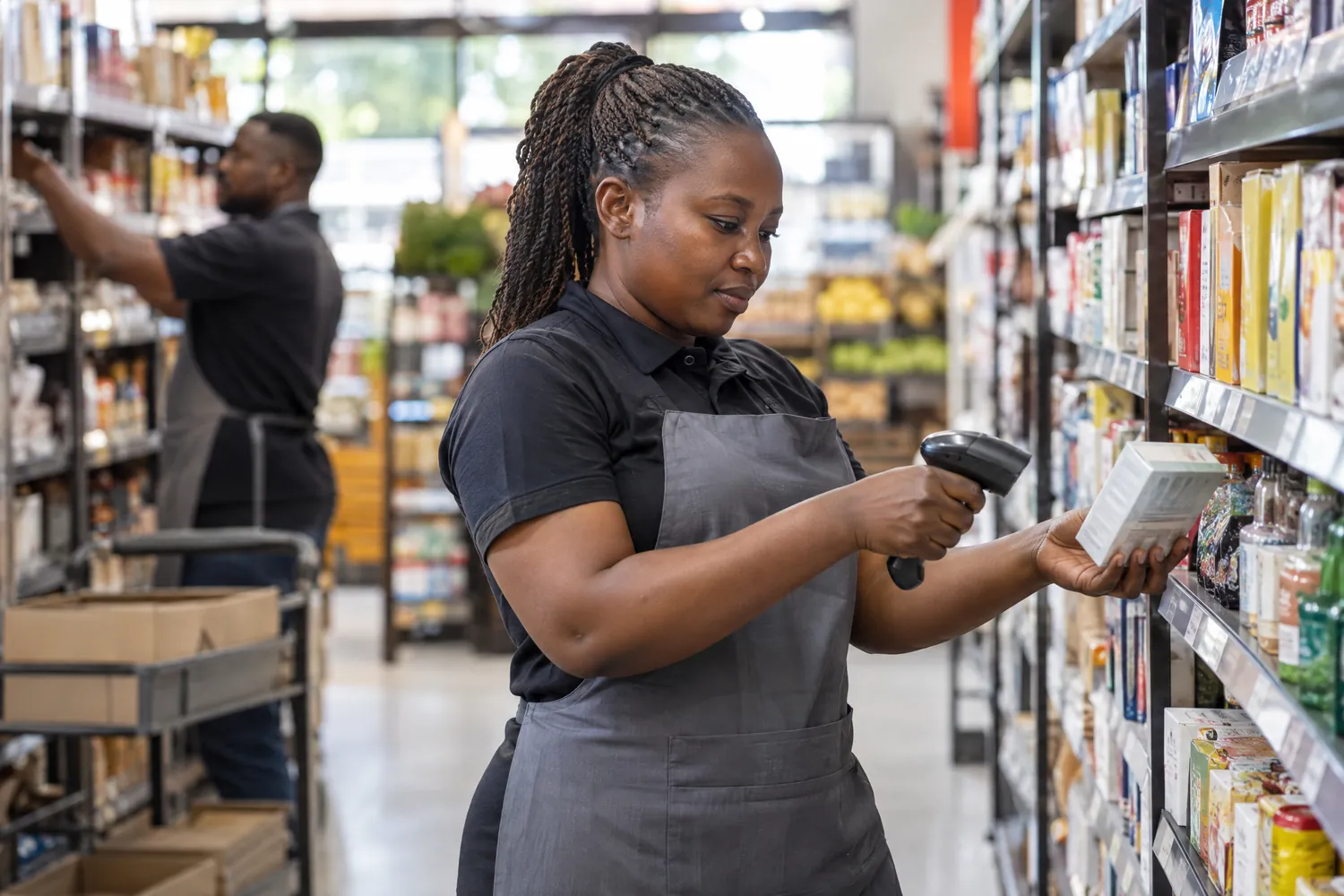 A store attendant scans stock on a shelf with a handheld barcode scanner while a colleague restocks behind her.
