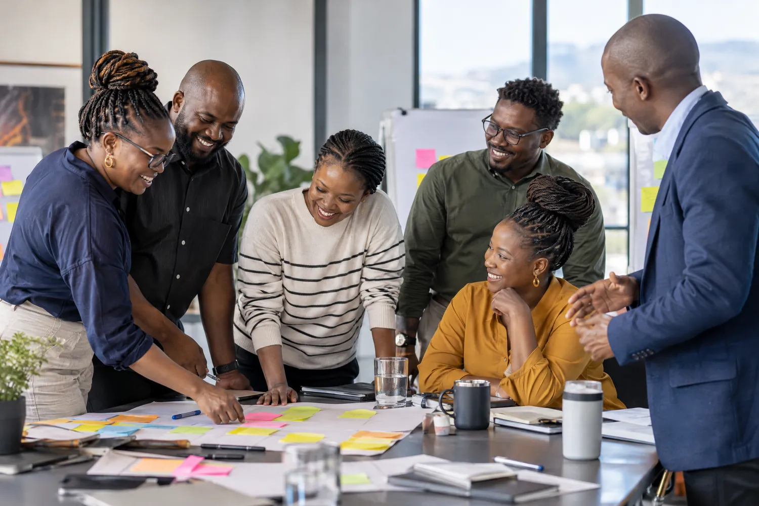 A team of colleagues laughs and collaborates over sticky notes spread across a workshop table.